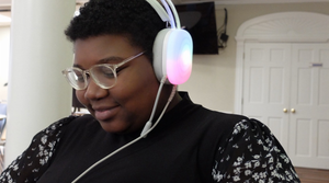 Woman smiling down at phone wearing clear rimmed glasses, white over the ear light up headphones, and black shirt with white flowers on the sleeves